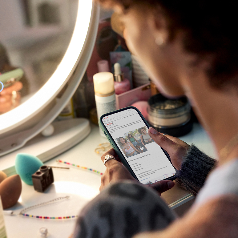 A woman sits in front of a mirror while holding her phone open to the  Here for Women web page on CVS.com