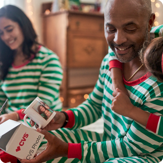 Family in matching green and white striped Christmas pajamas sitting around the tree with the father opening a customized mug from CVS Photo that has a family portrait on it while his daughter gives him a hug.