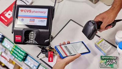 A bird’s eye view looking down at a point of sale transaction with a person holding a smartphone displaying a barcode that’s being scanned by the cashier.