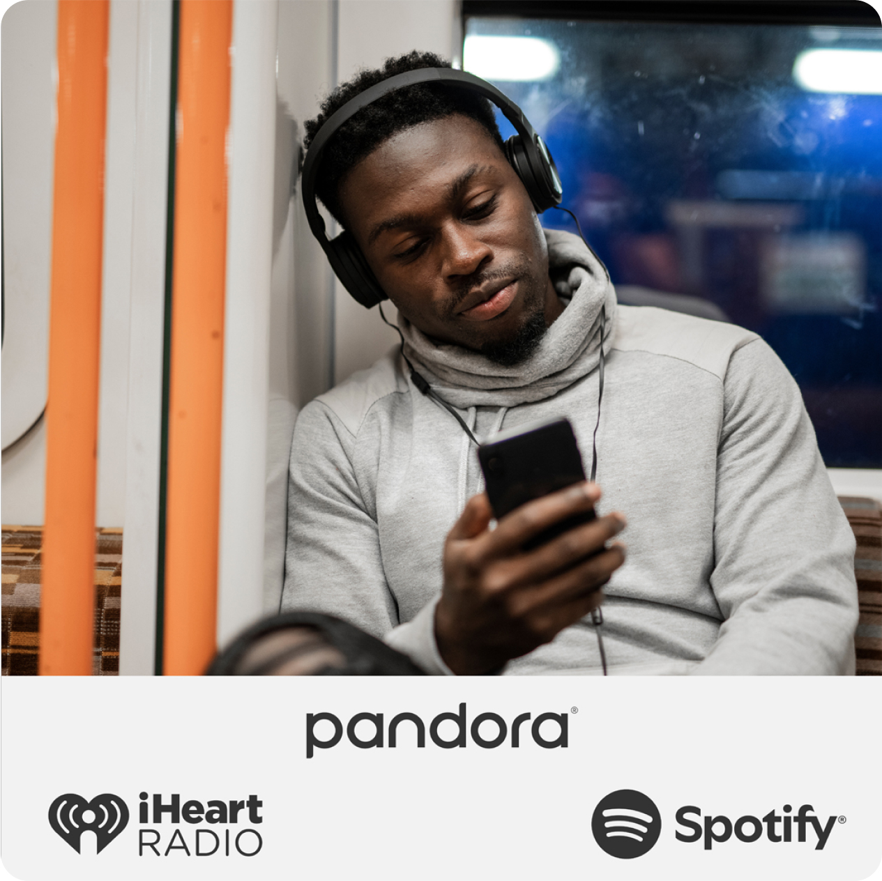 Relaxed man wearing headphones over his ears on the subway looking at his smartphone. The logos for Pandora, iHeart Radio and Spotify appear below.