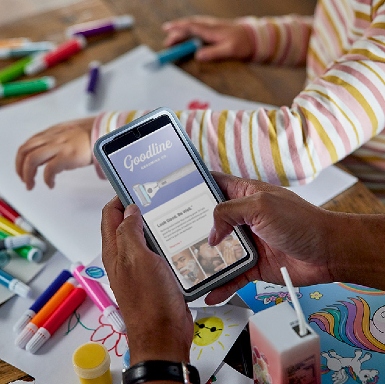 A child is coloring in the background while an adult scrolls through a Goodline Grooming Co. webpage on their smartphone.